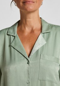 Chemise en satin vert clair avec un col, une poche plaquée et une fermeture à boutons. Le tissu a une texture lisse et brillante.
