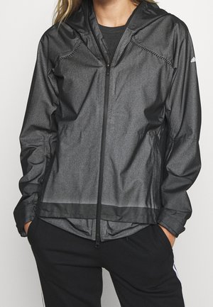 Person trägt eine schwarze Kapuzenjacke mit Reißverschluss, die Hände in den Taschen einer schwarzen Hose und zeigt das Adidas-Logo am linken Ärmel.