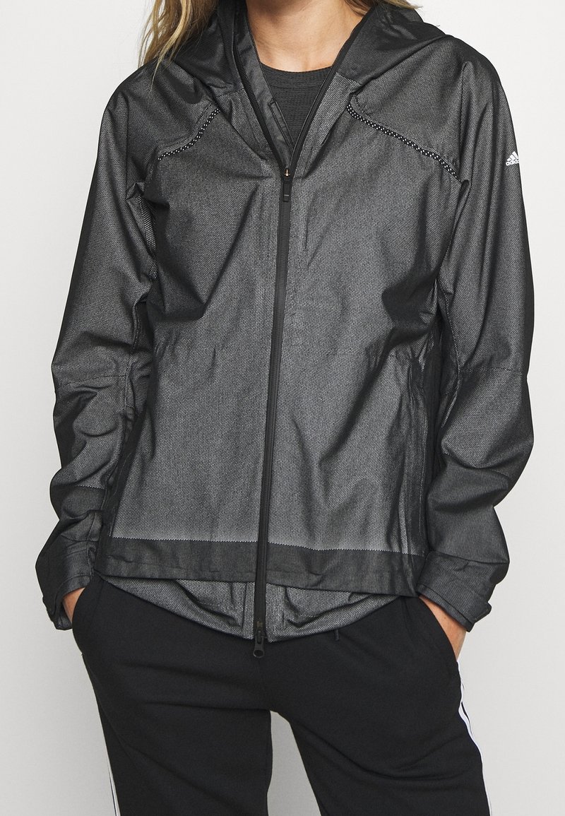 Personne portant une veste à capuche noire zippée, les mains dans les poches d’un pantalon noir, montrant le logo Adidas sur la manche gauche.