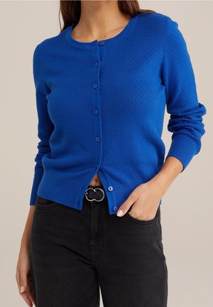 Femme portant un cardigan bleu texturé à boutons avec un pantalon noir, une main dans la poche et une ceinture noire à boucle ronde argentée.