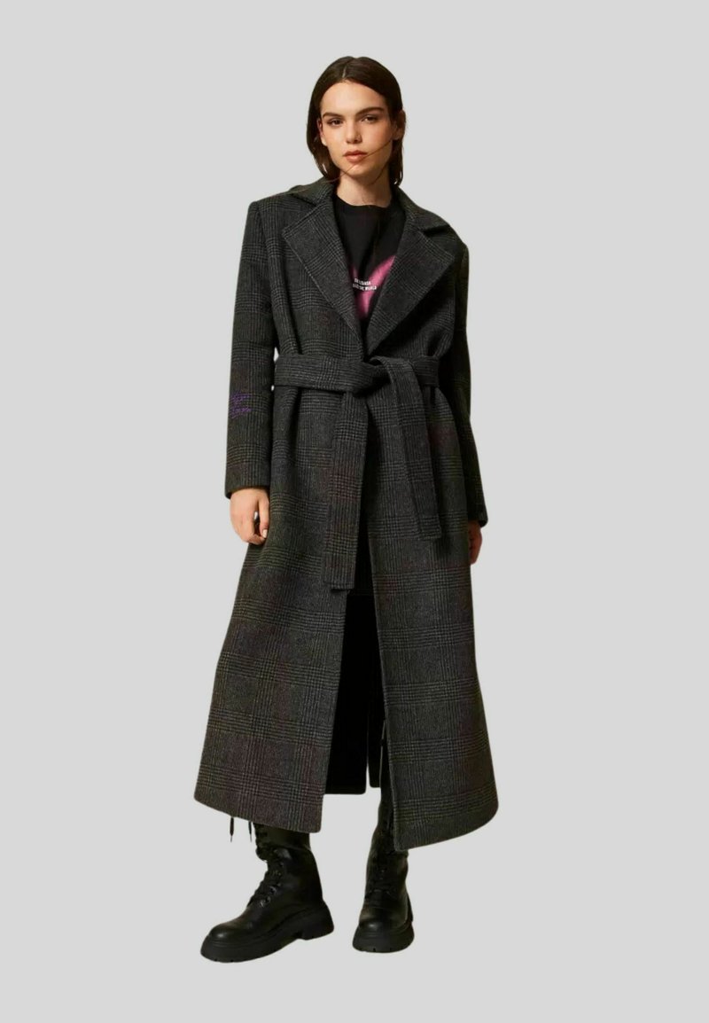 Cappotto lungo a quadri grigio scuro, con vita regolabile, colletto ampio e spacchi laterali. Indossato sopra una t-shirt grafica e abbinato a stivali neri.