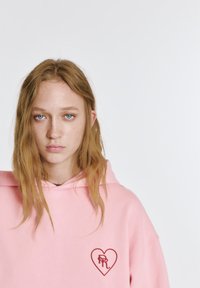 Ružová mikina s predným vreckom a červeným srdcovým logom s iniciálmi. Mäkká látka s voľným strihom a kapucňou so sťahovacou šnúrkou.