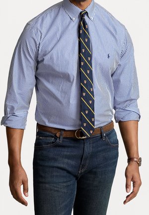 Homme portant une chemise rayée bleue et blanche boutonnée avec manches retroussées, cravate bleu marine à motif de blason, ceinture marron, jean foncé et montre marron.