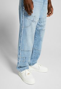 Vaaleansiniset farkut, joissa on repsottava sivusauma, useita taskuja ja rento istuvuus, yhdistettynä valkoisiin nilkkureihin, joissa on kultaiset yksityiskohdat.