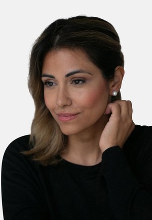 Nahaufnahme einer Frau mit dunklem Haar und Perlenohrringen, die ein schwarzes Langarmoberteil trägt, mit sanften, natürlichen Hauttönen und dezentem Make-up.