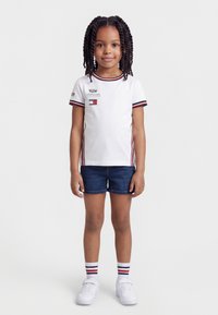 Νεαρή κοπέλα με πλεξούδες, φορώντας λευκό μπλουζάκι με λογότυπο, τζιν σορτς, ριγέ κάλτσες και λευκά αθλητικά παπούτσια, στέκεται μπροστά σε ένα απλό φόντο.