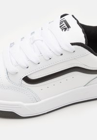 Baskets blanches avec une tige en cuir lisse, des accents noirs, des lacets blancs plats et des détails perforés sur les côtés pour la respirabilité.