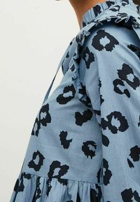 Vaaleansininen mekko mustalla seeprahraidalla. Siinä on röyhelökaulus ja hihat, liehuva helma ja se on valmistettu pehmeästä kankaasta.