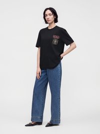 Γυναίκα με μαύρο T-shirt με στολίδια, φαρδιά μπλε τζιν παντελόνια και μαύρα μυτερά παπούτσια, στέκεται μπροστά σε ένα απλό φωτεινό φόντο.