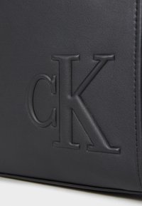 Black leather bag featuring embossed "CK" logo. Smooth texture with detailed stitching along edges. Rectangular shape with minimalist design.