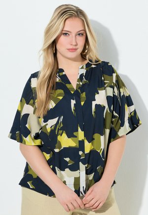 Frau mit langem blondem Haar, die goldene Creolenohrringe trägt und eine lockere Kurzarmbluse mit einem abstrakten Blumendruck in Marineblau, Grün, Gelb und Weiß.