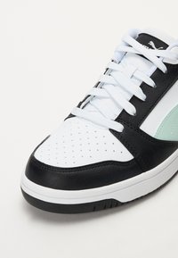 Detailní záběr na černou, bílou a mentolově zelenou tenisku s bílými tkaničkami a perforovanou špičkou na bílém pozadí.