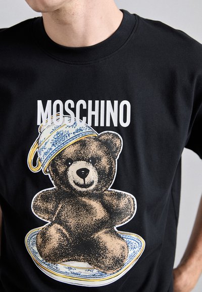T-shirt noir avec un graphique d'un ours brun dans une soucoupe à motifs bleus portant un chapeau bleu et jaune, avec "MOSCHINO" imprimé au-dessus.
