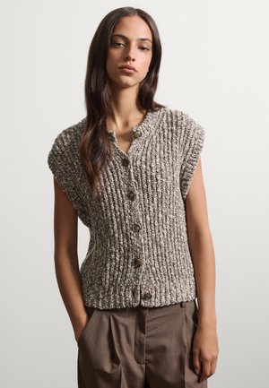 Jeune femme aux longs cheveux foncés portant un gilet tricoté marron sans manches boutonné et un pantalon marron ample, les mains dans les poches.