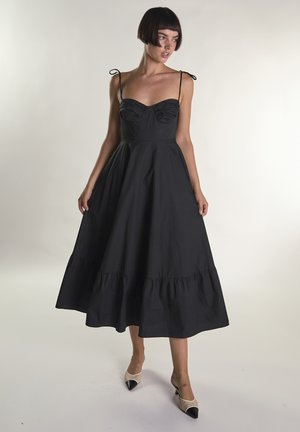 Kobieta ubrana w czarną sukienkę midi bez rękawów z wiązanymi ramiączkami i dopasowanym gorsetem, połączona z beżowymi, szpiczastymi butami z czarnymi czubkami.