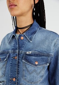 Jasnoniebieska kurtka dżinsowa z miedzianymi guzikami, kieszeniami na piersi z ozdobnym szwem oraz z metką marki na lewej kieszeni.