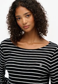 Top de rayas blancas y negras de manga larga con un escote amplio y un pequeño logo bordado en el pecho. Tejido suave y texturizado.