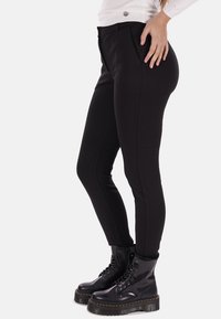 Pantaloni neri slim-fit con texture liscia, dotati di tasche laterali e una sottile piega frontale. Abbinati a stivali da combattimento neri con suola spessa.