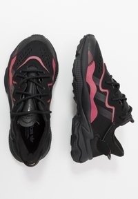 Černé sportovní boty mají vršek z pružné síťoviny s růžovými akcenty, texturovanou podrážku a tradiční tkaničky. Výrazným prvkem designu jsou vrstvené materiály.