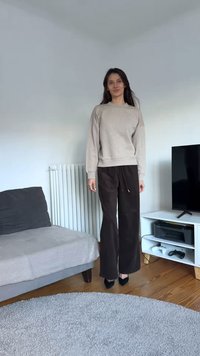 Sweatshirt cinzento claro, calças largas castanho escuro, sapatos pretos de salto alto, em pé sobre pavimento em madeira. Tapete cinzento suave junto a um sofá e uma TV.