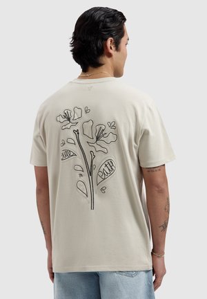 Man draagt een licht beige T-shirt met zwart omlijnd bloemenpatroon en de woorden "liefde" en "geloof" op de achterkant, gecombineerd met lichtblauwe spijkerbroek.