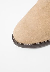 Stivale in suede beige con punta arrotondata, caratterizzato da una suola scura a contrasto e cuciture visibili lungo il bordo. Texture complessivamente liscia.