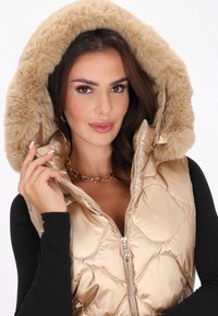 Vesta metalizată aurie matlasată cu glugă din blană artificială, având un fermoar frontal și un design ajustat, purtată peste un top negru cu mânecă lungă.