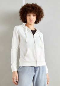 Bijela zip-up dukserica napravljena od mekog materijala, ima kapuljaču, prednje dzepove i rebraste manžetne. Uparena s svijetloplavim farmerkama.