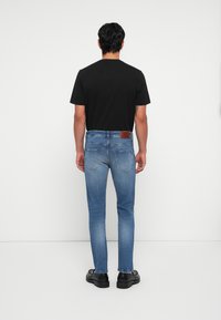 Man wearing black t-shirt, slim light blue jeans, and black shoes, standing facing a white wall with arms relaxed by sides.