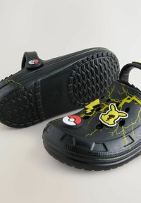 Zwarte Crocs met een gestructureerde rubberen zool en drie decoratieve elementen: een Poké Ball, Pikachu en gele bliksemschichten.