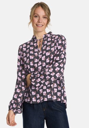 Frau lächelt und trägt eine langärmlige schwarze Bluse mit pinken Blumenmustern und dunkelblauen Jeans, während sie mit einem Arm über ihren Körper posiert.