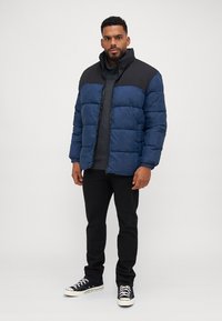 Doudoune marine avec épaules noires, fermeture éclair et panneaux cousus ; portée sur un pull gris et un pantalon noir, associée à des baskets.