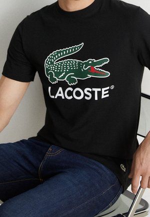 Schwarzes Baumwoll-T-Shirt mit einem grünen Krokodil-Logo und weißem "LACOSTE"-Schriftzug. Kombiniert mit blauen Jeans, sitzend auf einem Stuhl dargestellt.