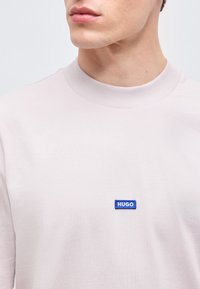 Κοντινό πλάνο ενός ατόμου που φοράει φανέλα ανοιχτό ροζ με στρογγυλή λαιμόκοψη και μικρό μπλε ορθογώνιο λογότυπο "HUGO" στο στήθος.