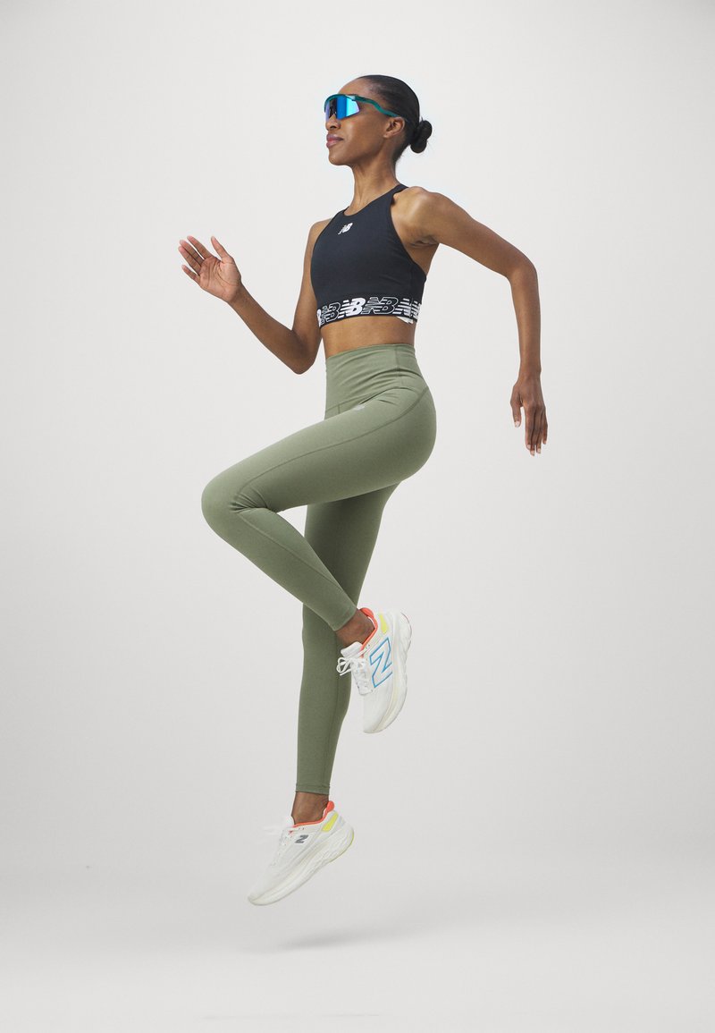 Top deportivo negro corto con una banda blanca de marca, leggings verde olivo y zapatillas blancas con acentos coloridos, posando en medio de un salto.