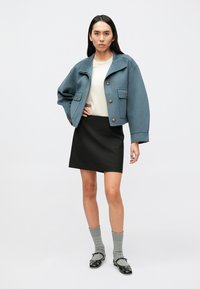 Τζάκετ σε τιρκουάζ χρώμα, κοντό, με χρυσά κουμπιά, συνδυασμένο με εκρού μπλούζα και μαύρη μίνι φούστα, συμπληρωμένο με γκρι κάλτσες και μαύρα φλατ παπούτσια.