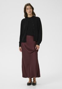 Schwarze Strickjacke über einem schwarzen Oberteil, kombiniert mit einem langen burgunderfarbenen Satinrock. Das Model trägt schwarze flache Schuhe, die eine glatte Textur und ein schlichtes Design aufweisen.