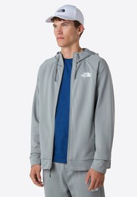 Lys grå zip-up jakke med et tekstureret mønster, fastgjort hætte og to frontlommer. Bæres over en blå mønstret skjorte og grå bukser.