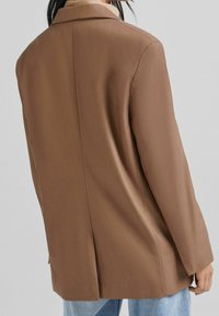 Hnědý blejzr s hladkou texturou, s jedním rozparkem na zádech a výřezovými klopami, kombinovaný se světle modrými džínami.