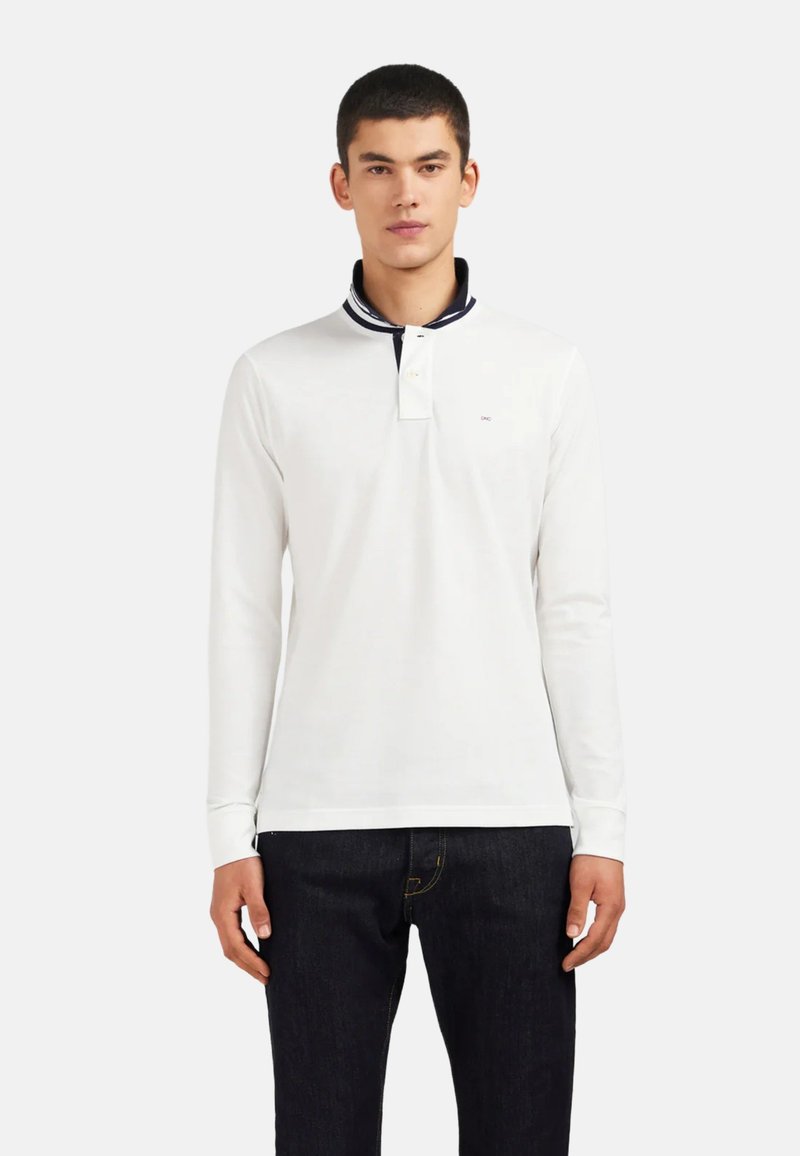 Jeune homme portant un polo blanc à manches longues avec un col rayé bleu marine et un jean foncé, se tenant contre un fond clair unicolore.
