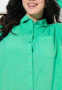 Chemise verte à boutons en tissu léger, dotée d'une poche sur le côté gauche et d'une patte de boutonnage à l'avant avec des boutons assortis.
