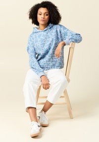 Felpa con motivo floreale blu e coulisse, abbinata a pantaloni cropped bianchi e sneakers bianche con accenti neri, seduta su una sedia di legno.