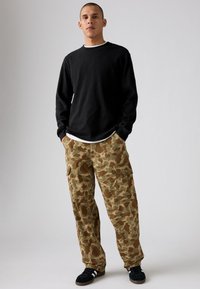 Camisa de manga larga negra con cuello color crema, combinada con pantalones cargo de camuflaje que cuentan con múltiples bolsillos en tonos tierra, llevados con zapatillas negras.
