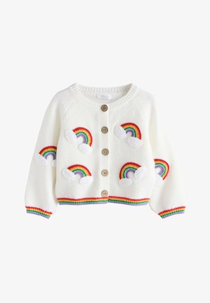 Kleines Kind mit dunklen Haaren im Dutt, trägt eine weiße Strickjacke mit Regenbogenaufnähern, weite Jeanshosen mit Rüschensäumen und weiße Schuhe.