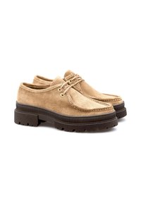 Une paire de chaussures à lacets en daim beige avec des semelles épaisses en caoutchouc noir et des détails de couture, présentée sur un fond blanc.
