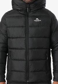 Veste matelassée noire avec capuche, fermeture éclair à l'avant, poches zippées sur les côtés, et logo "Helvetica" blanc sur la poitrine gauche portée par une personne.