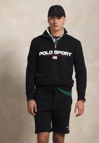 Fekete pulóver "POLO SPORT" felirattal, cipzáras nyakkal, zöld galléros ing felett viselve, fekete rövidnadrággal és fekete sapkával párosítva. A modell áll.