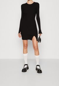 Robe noire à manches longues côtelée avec une fente latérale, dotée d'un col rond et d'une silhouette ajustée, associée à des chaussettes blanches et des chaussures noires épaisses.