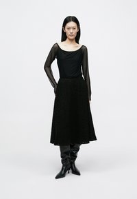 Sort langærmet top i gennemsigtigt stof, kombineret med et knælange tekstureret sort nederdel. Iført knælange sorte støvler med hæl.