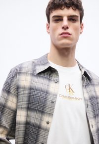 Νεαρός άνδρας που φοράει ένα πουκάμισο με γκρι και λευκό καρό κουμπωμένο πάνω από ένα λευκό t-shirt Calvin Klein Jeans με λογότυπο.